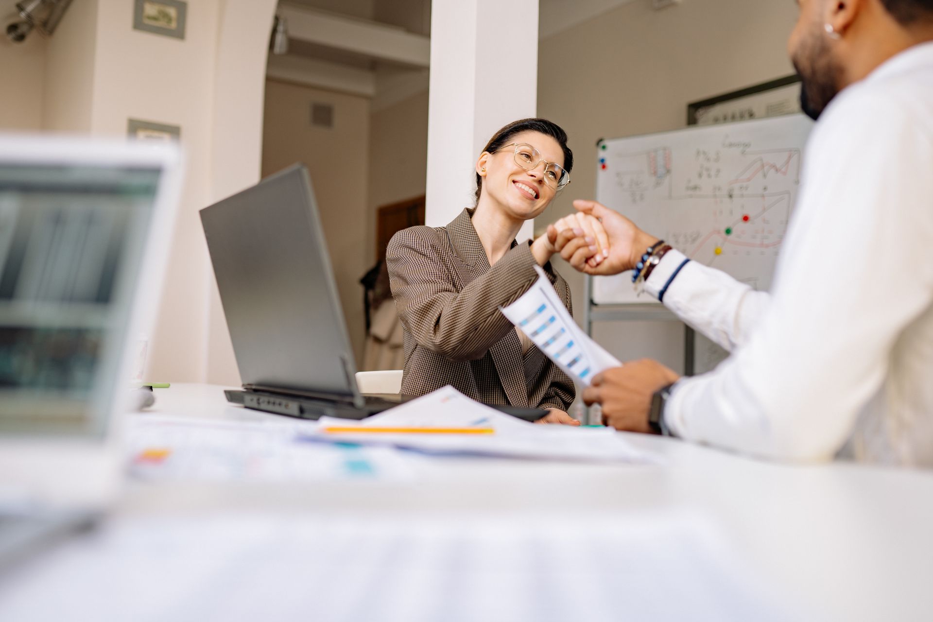 Professional woman in a blazer shaking hands with a sales candidate across a desk after a successful interview