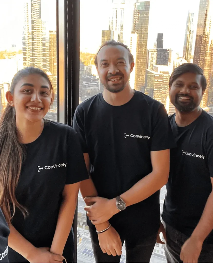 Three people wearing black Convincely t-shirts stand together and smile in front of large windows with a city skyline in the background.