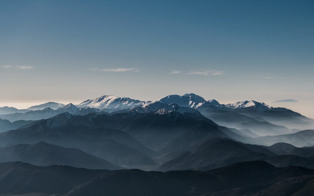 A range of mountains with snow-covered peaks in the distance under a clear blue sky, with mist settling in the valleys.