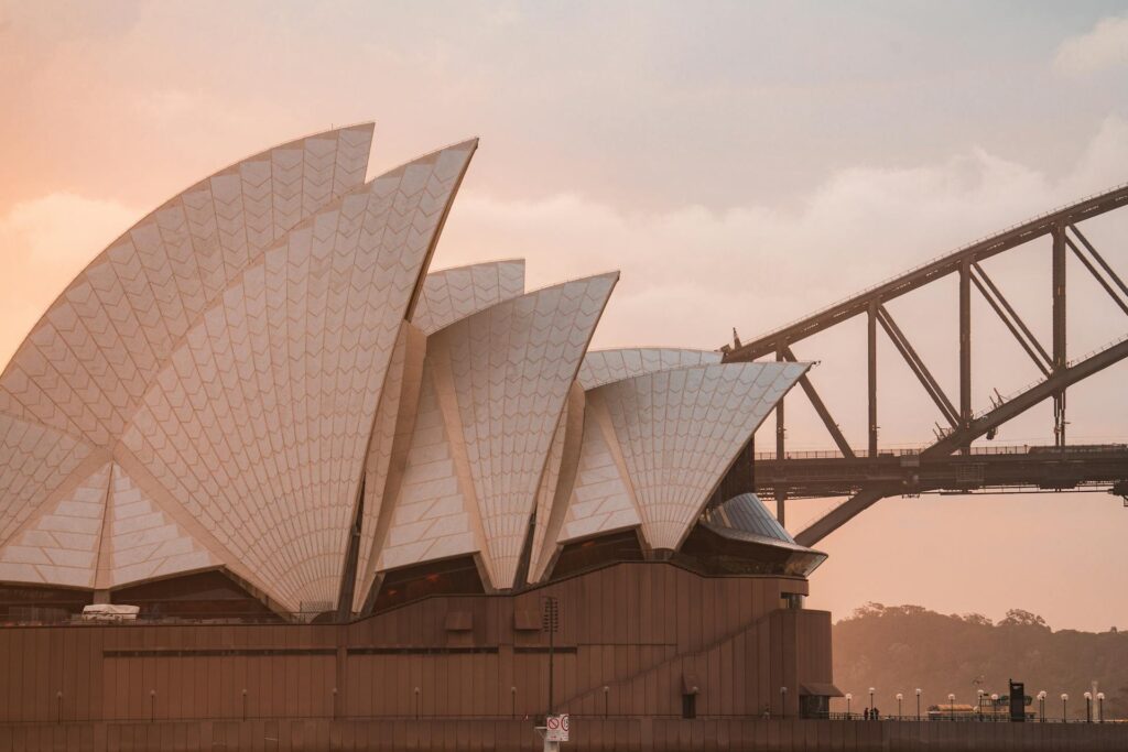 Sydney Opera House with its white sail-like roofs in the foreground, and part of the Sydney Harbour Bridge visible in the background at sunset.