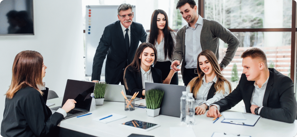 Six professionals in business attire have a discussion around a conference table with laptops, notebooks, and water bottles in a modern office setting.