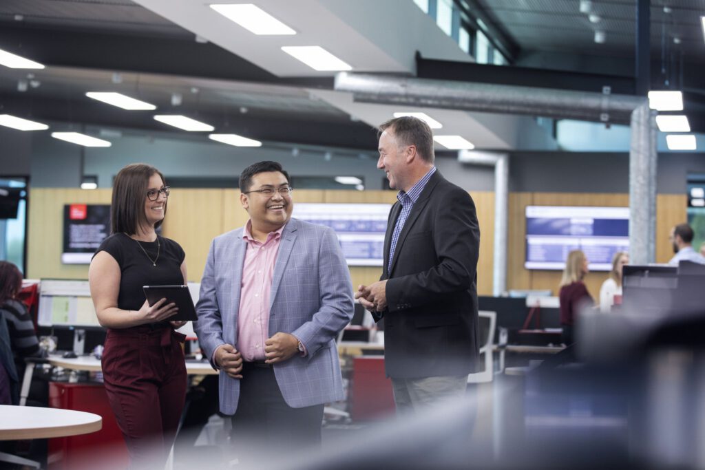 Three people in business attire stand and converse in a modern office with desks, computers, and large digital screens in the background.