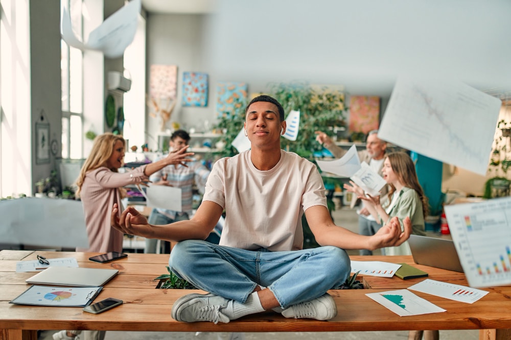 A young man sits cross-legged on a desk meditating calmly while coworkers argue and papers fly around in a chaotic office environment.