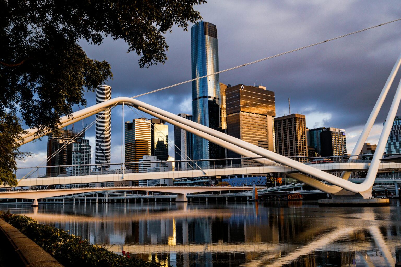 A modern white footbridge spans over a calm river with tall city skyscrapers and reflections visible in the background during daylight, illustrating the dynamic growth seen in areas where a Sales Recruitment Agency Brisbane might thrive.