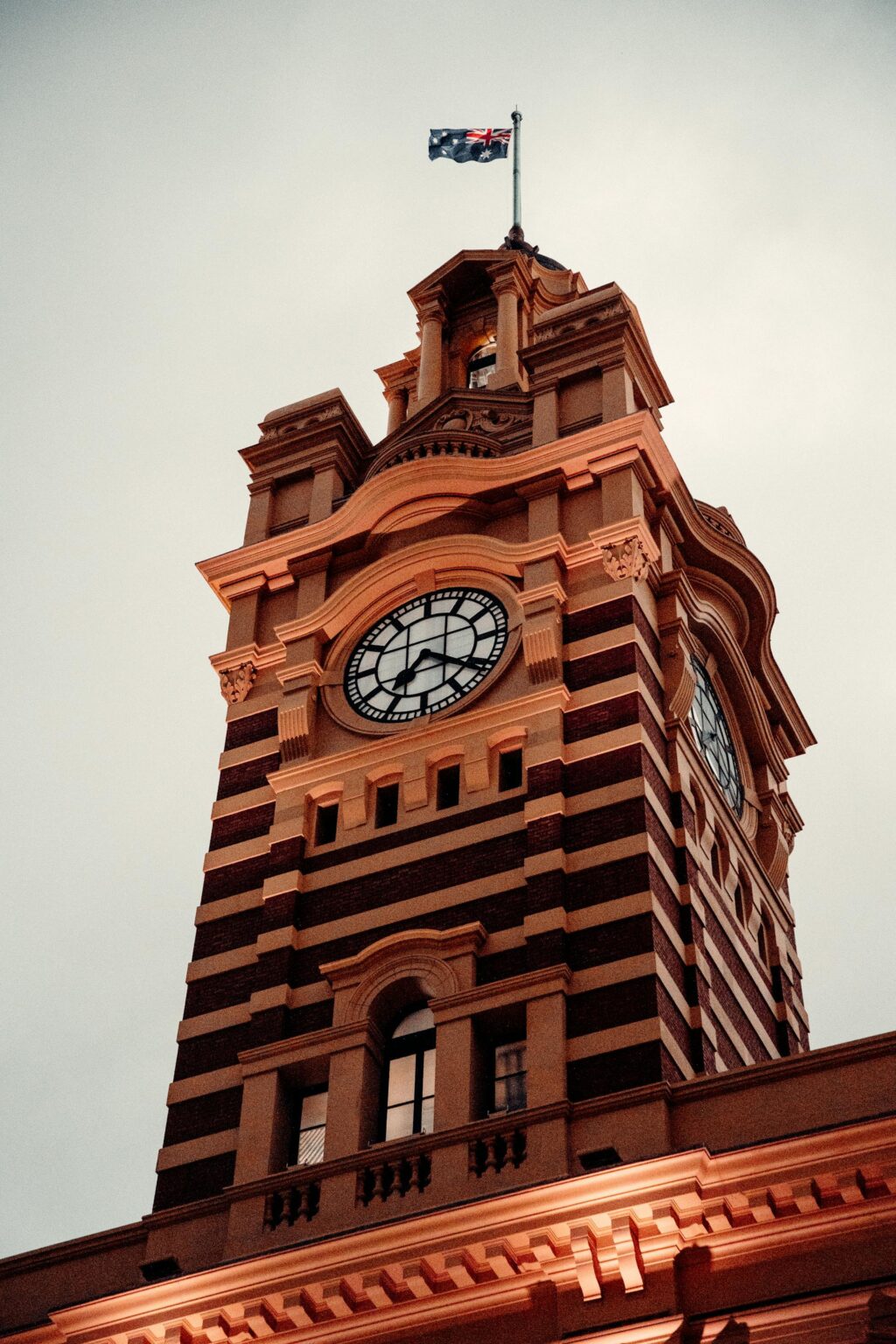 A tall brick clock tower with a striped pattern, flying an Australian flag, stands against a cloudy sky—a striking sight in the heart of Melbourne, near leading Sales Recruitment Agency Melbourne offices.