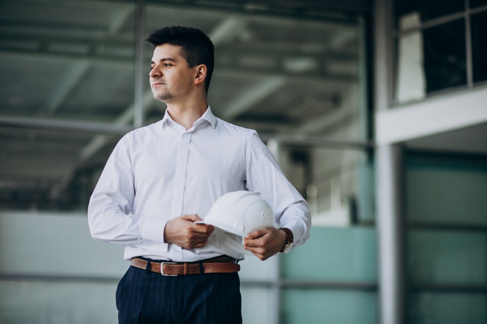 A man in a white dress shirt holding a white safety helmet stands indoors, looking to the side.