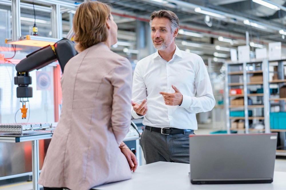 Two people in business attire talk in an industrial workspace near a robotic arm and a laptop, with shelves and equipment in the background.