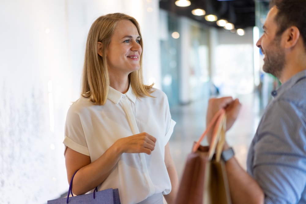 A woman and a man stand indoors, holding shopping bags and having a conversation.