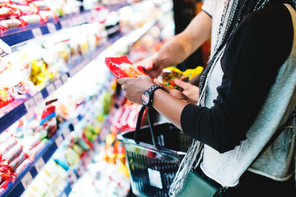 Two people stand in front of a brightly lit grocery store shelf, holding packaged snacks and a shopping basket.