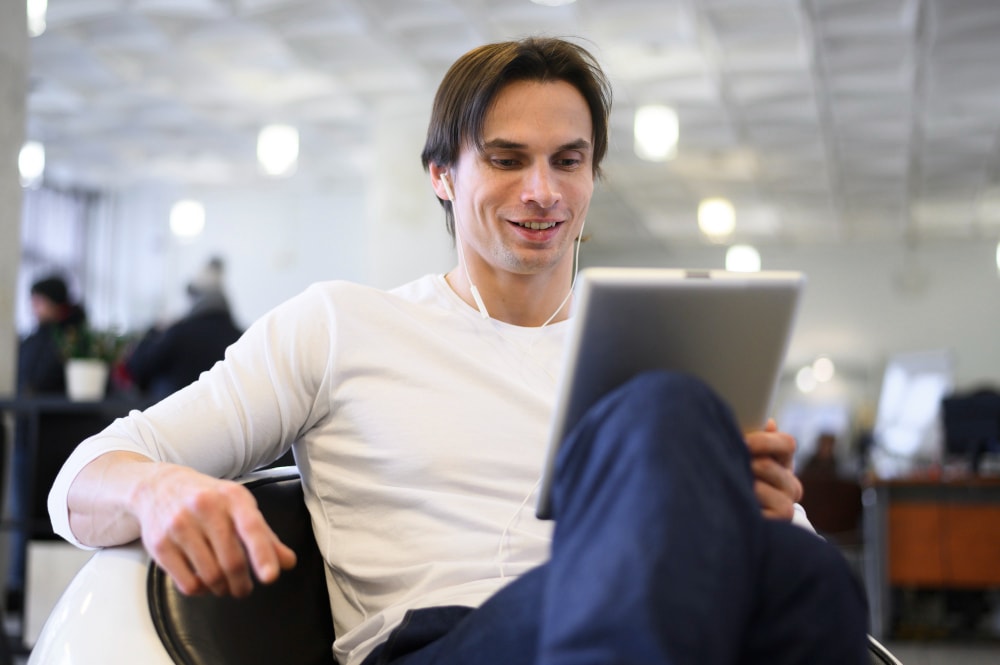 A man wearing a white long-sleeve shirt sits in a chair, using a tablet and listening with wired earphones in a bright, modern indoor space.