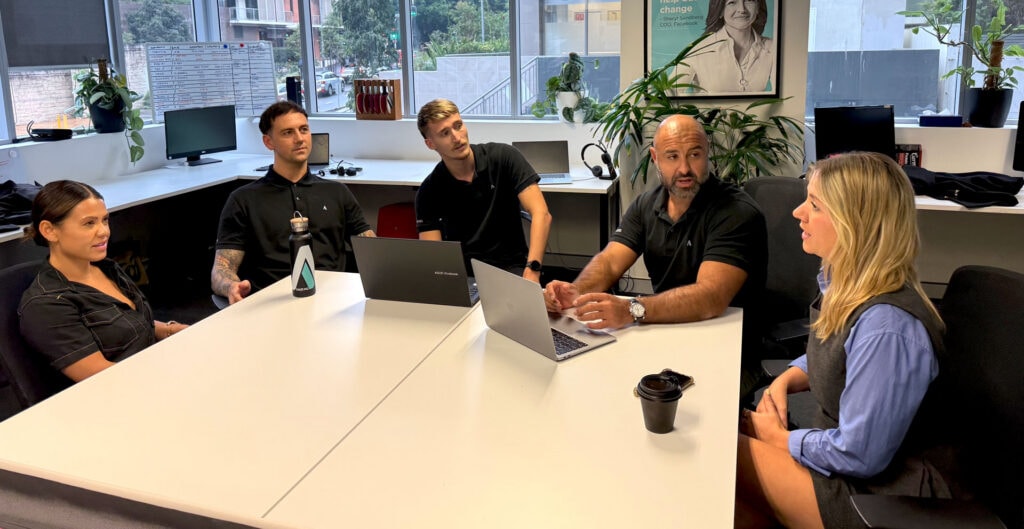 Five people sit around a desk in an office engaged in a discussion, with laptops, a water bottle, and a coffee cup on the table.