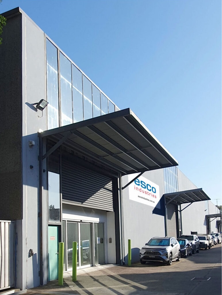 Exterior view of a modern industrial building with an esco industries sign, a large roller door, parked cars, and glass windows along the upper facade.
