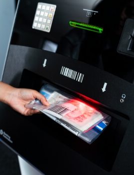 A hand is inserting an envelope with cash under a barcode scanner at an automated deposit machine.