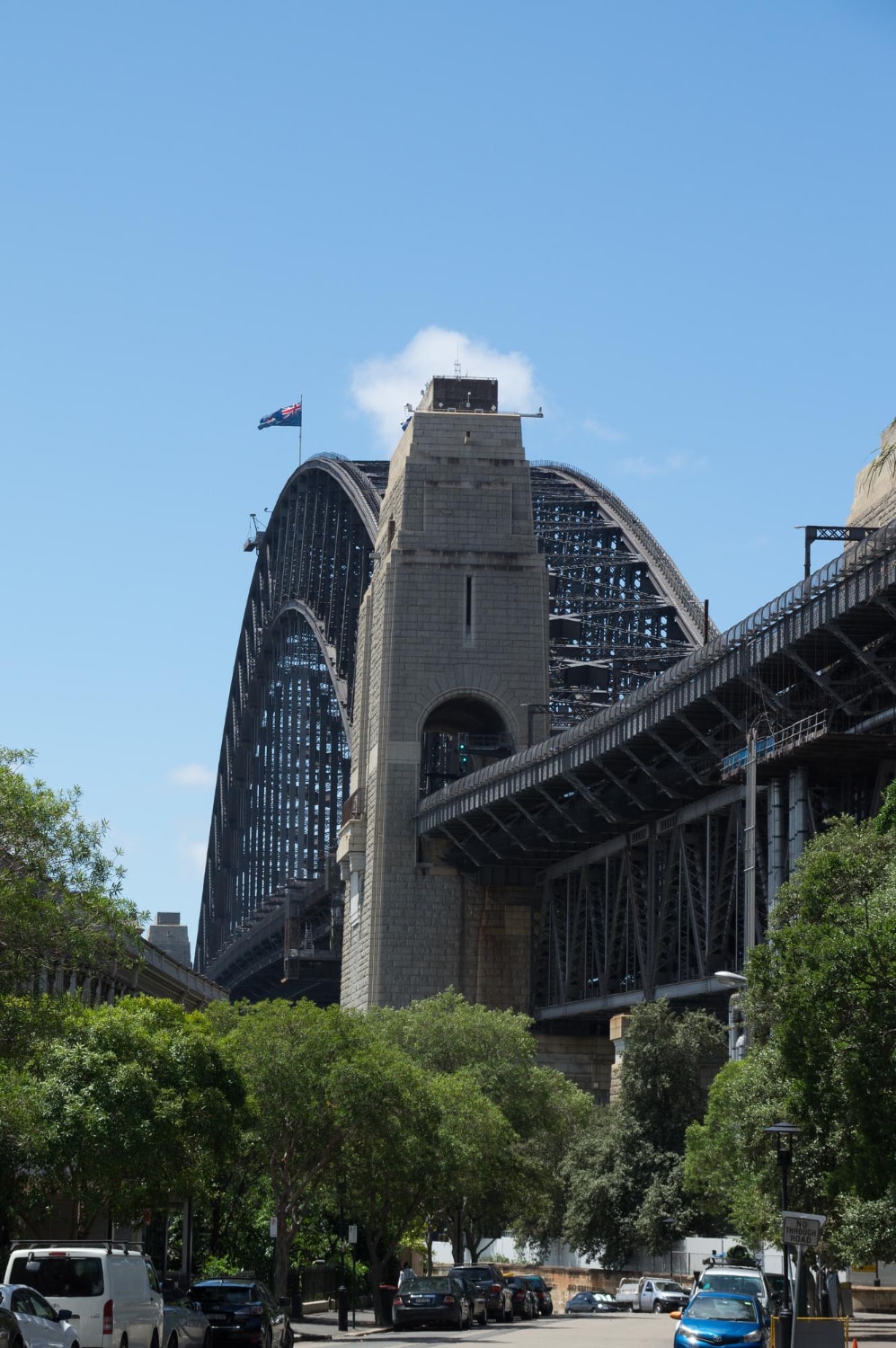 Sydney Harbour Bridge with an Australian flag on top, viewed from below on a sunny day with cars parked along the street and trees in the foreground.