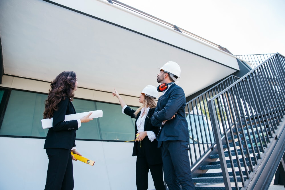 Three people in business attire and hard hats stand outside a modern building, discussing and examining architectural plans near a staircase.