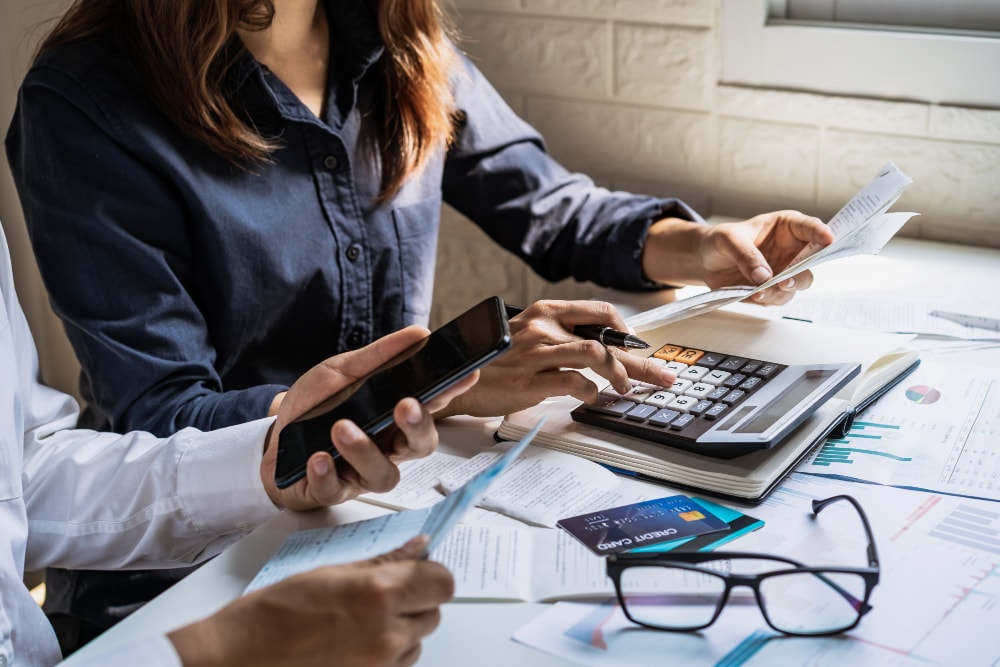 Two people review financial documents at a desk with a calculator, smartphone, credit card, and paperwork spread out, suggesting a budgeting or financial planning activity.