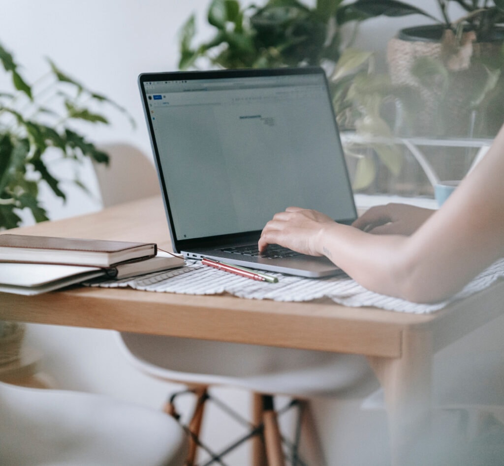 A person types on a laptop at a wooden table with notebooks, a pencil, and plants in the background.
