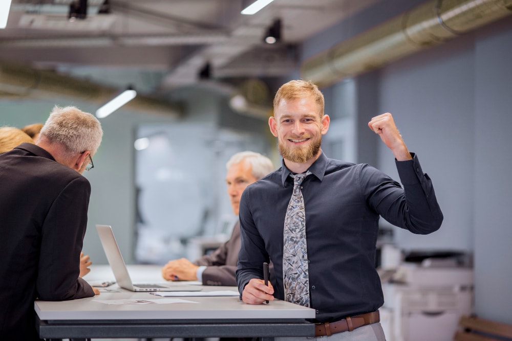 A man in business attire stands at a desk smiling and raising his fist, while three other people work in the background in an office setting.