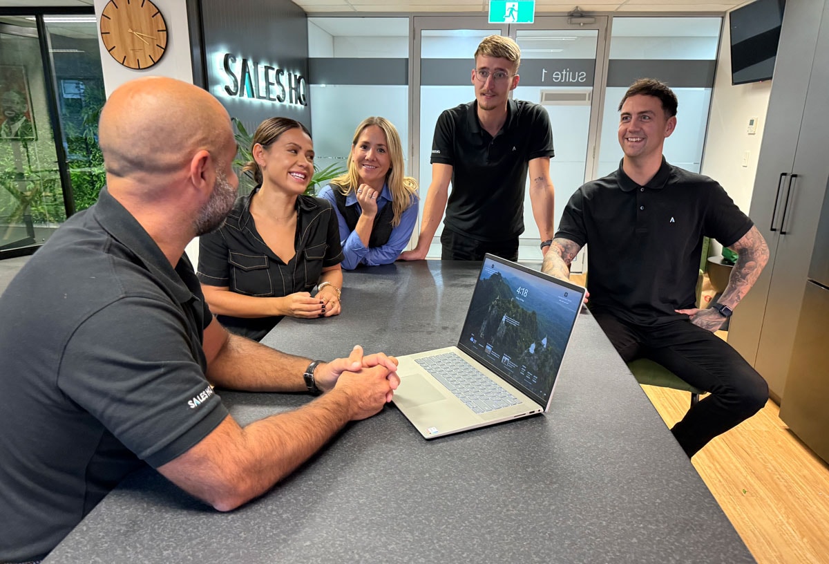 Four people stand around a table listening to a seated man with a laptop open in a modern office setting.