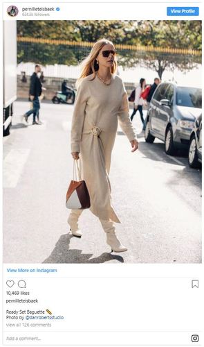 A woman wearing a beige sweater dress, white boots, and sunglasses walks on a city street carrying a brown and white handbag.