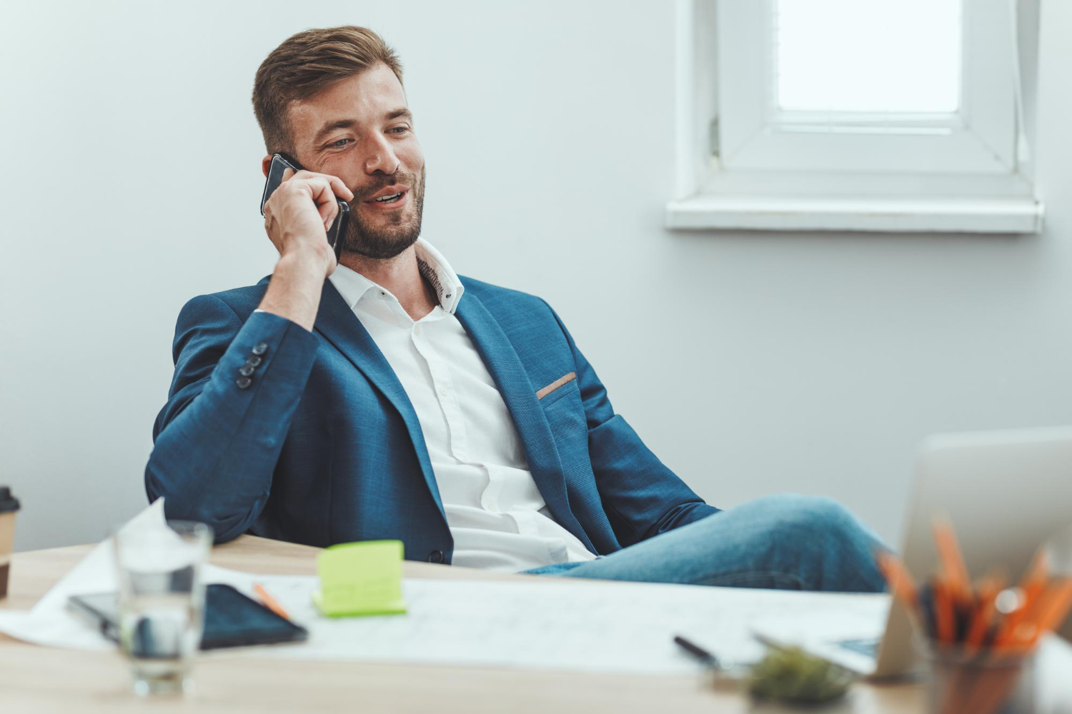 Man in a blue suit sitting at a desk, making sales calls on his smartphone, with papers, a laptop, and office supplies spread out before him.