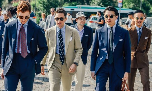 A group of five men wearing suits and sunglasses walk outdoors on a sunny day, with trees and a bus in the background.