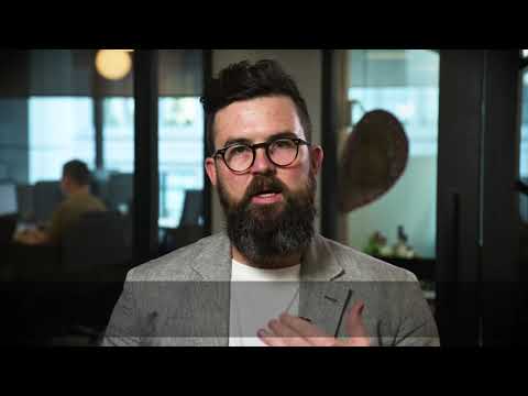A bearded man wearing glasses and a light blazer speaks in an office setting, with another person working in the background.